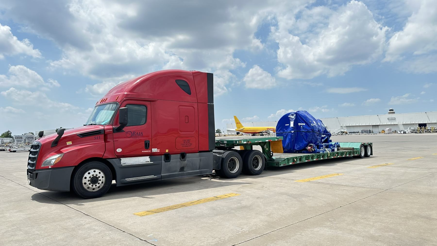 Red Raja Express truck with a covered Pratt & Whitney jet engine on an RGN at the Dallas air cargo ramp, a cargo jet behind it